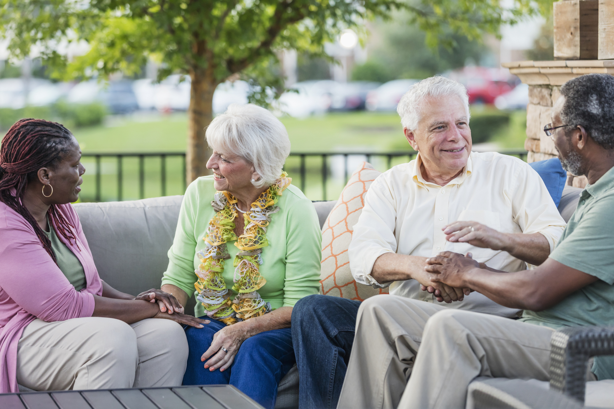 A multi-ethnic group of mature and senior friends hanging out together, sitting outdoors on a patio conversing. The African-American couple are in their 50s. The Caucasian couple are in their 70s. The women are talking and holding hands, and the men are chatting with each other.