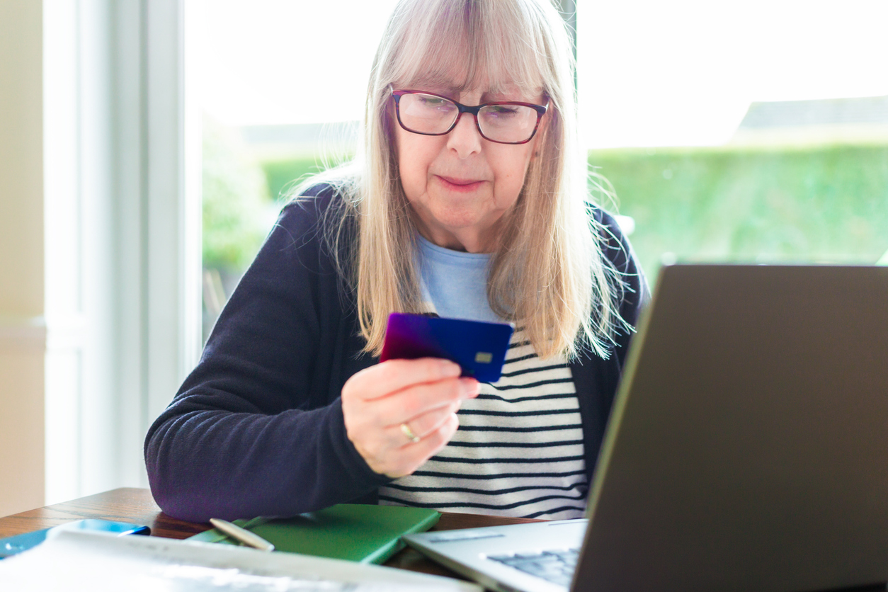 Portrait of a retired senior woman in her 70s making a payment online at home using her smart phone and laptop computer.