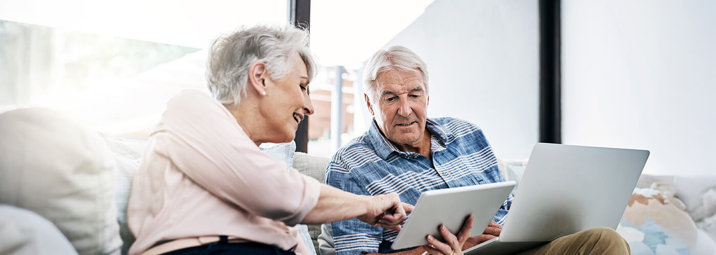 Senior man and woman sit on a couch and review information on a laptop and tablet screen together