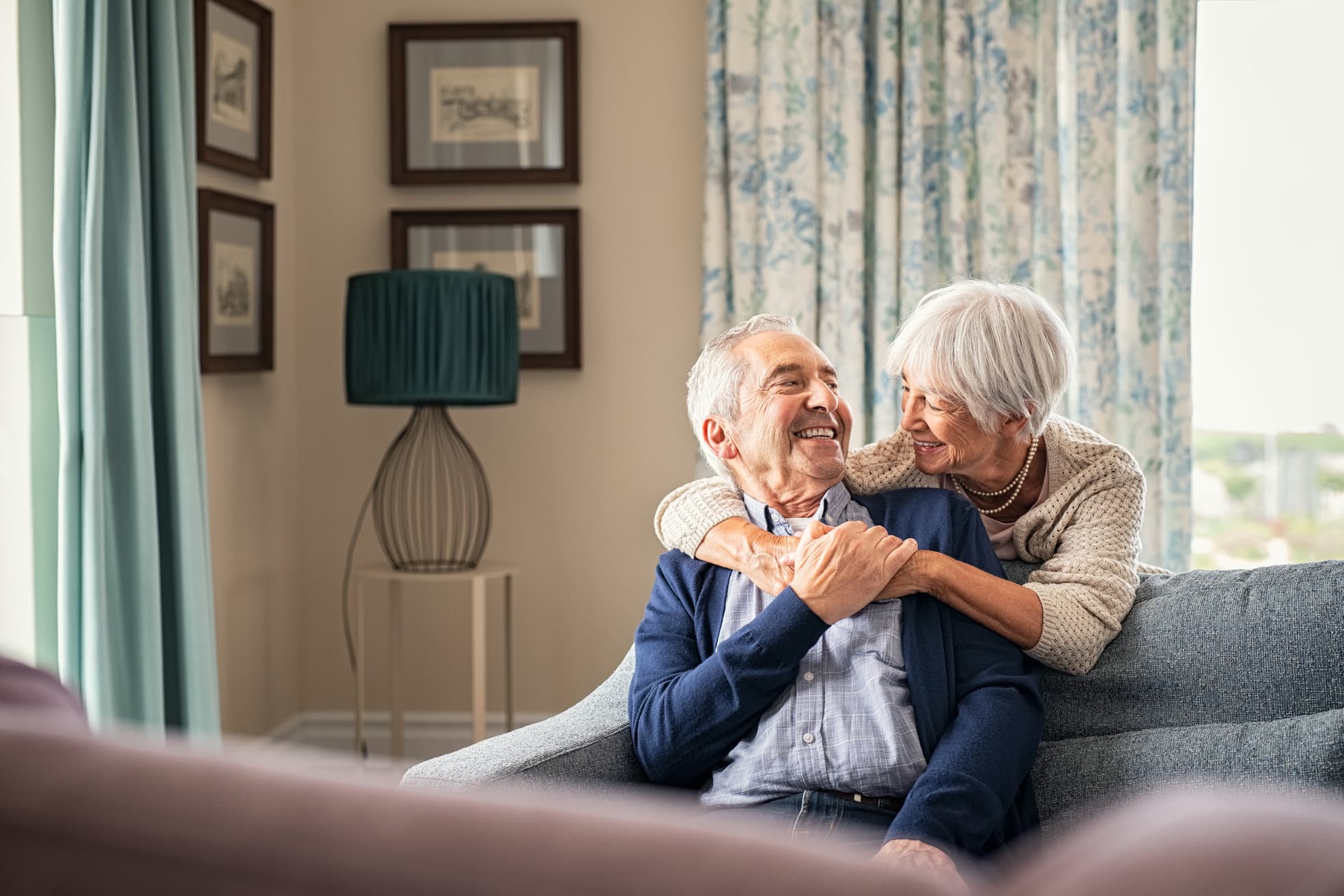 Senior woman stands behind couch and embraces senior man while they smile at each other